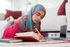 A female student wearing a hijab studies at home, using a notebook, pen and laptop