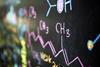 A close-up photograph of a chemical formula written in pink chalk on a blackboard, with other chemical structures drawn in different colours in the background